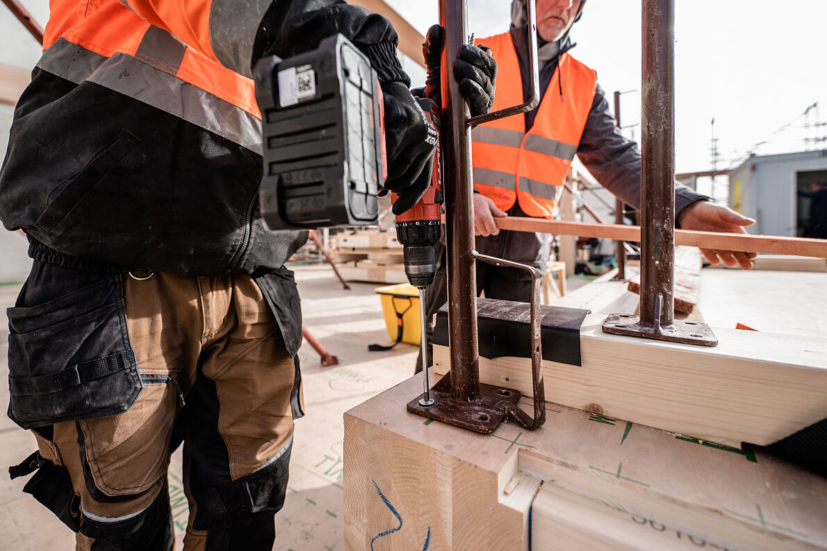 Handwerk und Präzision: Zwei Zimmerleute befestigen Balken, ein Schritt auf dem Weg, den TRI als Münchens Holz-Hybrid-Vorreiter zu vollenden.